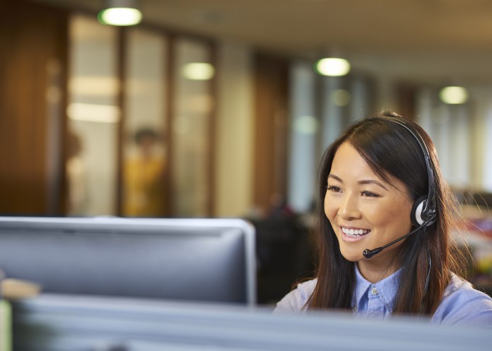 a young call centre representative greets a caller in a large open plan office . Co-workers can be seen defocussed in the background .